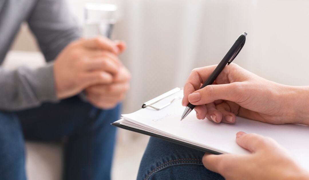 A close up of a person writing things down on a clipboard with another person across from them with their hands clasped.