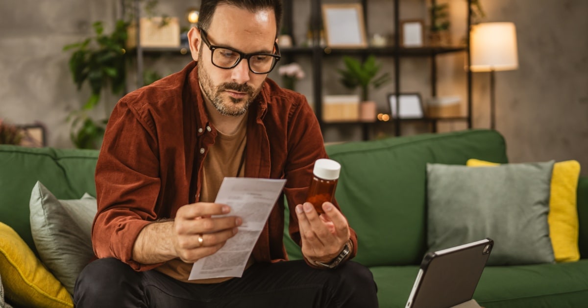 A person on a couch holds a pill bottle and instructions with more pills and an iPad on the coffee table in front of him.