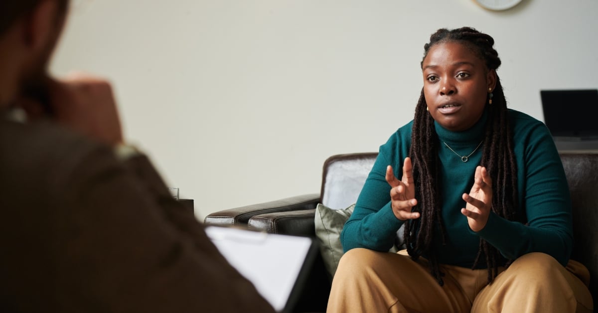 A young woman sits across from a psychologist in an office, gesturing with their hands with a clock behind them.