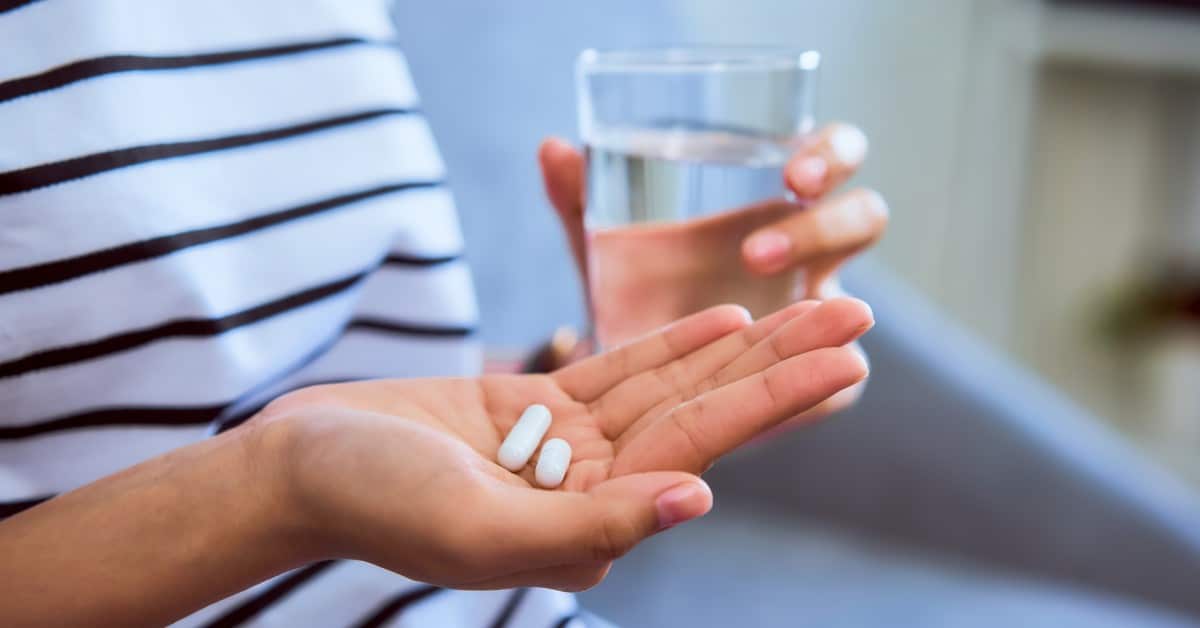 A person on a couch holds a glass of water in their left hand and two non-descript pills in their right hand.