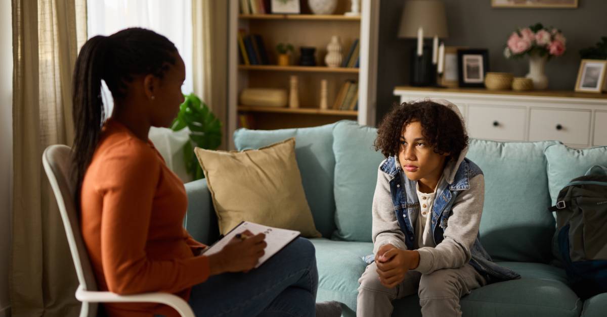 A child sits on a couch in a therapists office while a woman with a clipboard sits in a chair opposite the child.