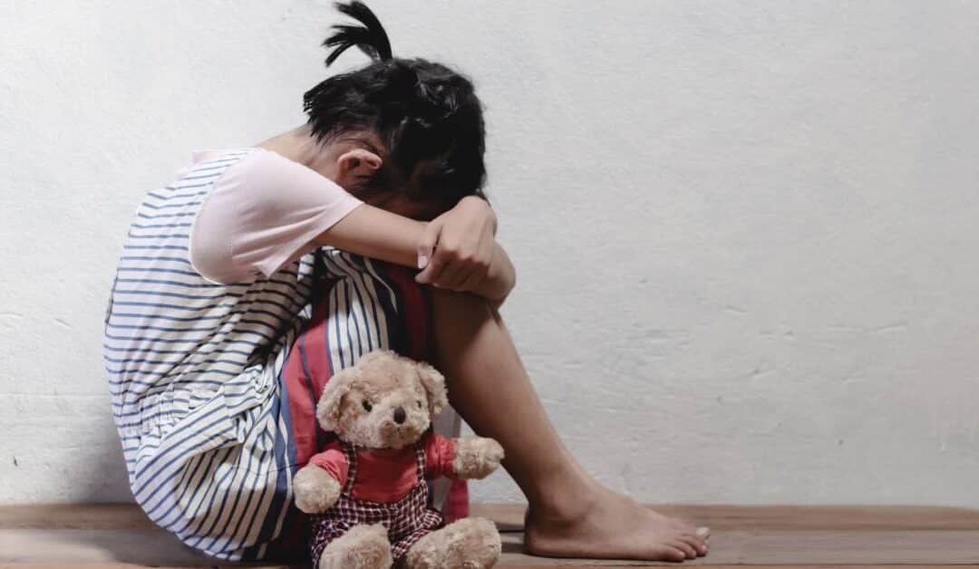 A kid is curled up in a corner of a room with their head in their knees and a small stuffed bear next to them.