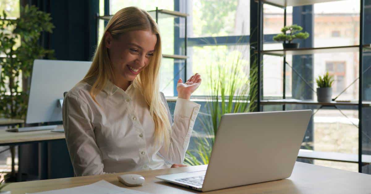 A woman looking at her laptop with a smile on her face. Potted plants sit on shelving in front of a window in the background.