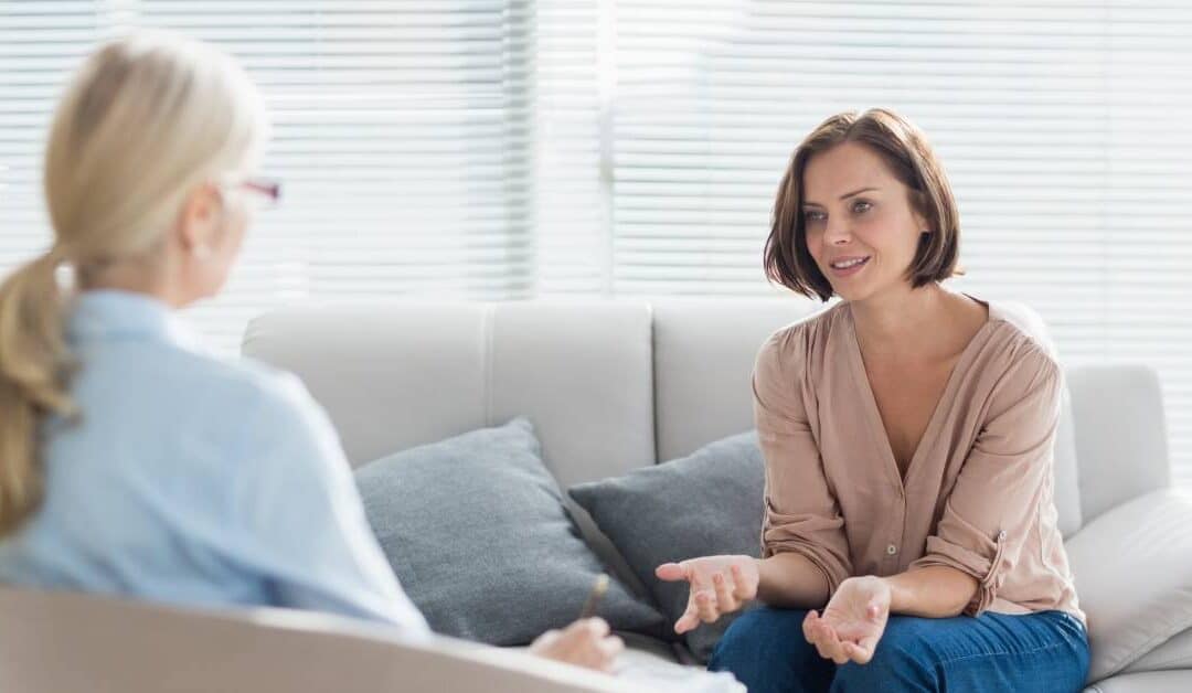 A woman sitting on a couch with pillows, gesturing with her hands at another woman in a chair opposite of her.