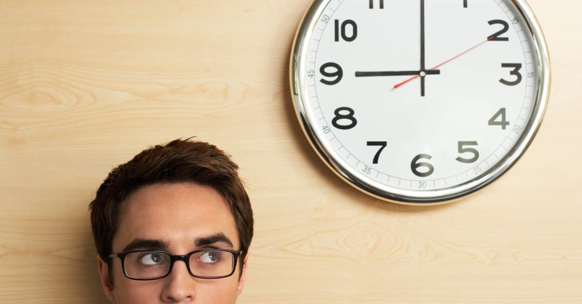 A man with brown hair and glasses looks at a clock on a wall that reads 9 o'clock. The wall features a tan color.