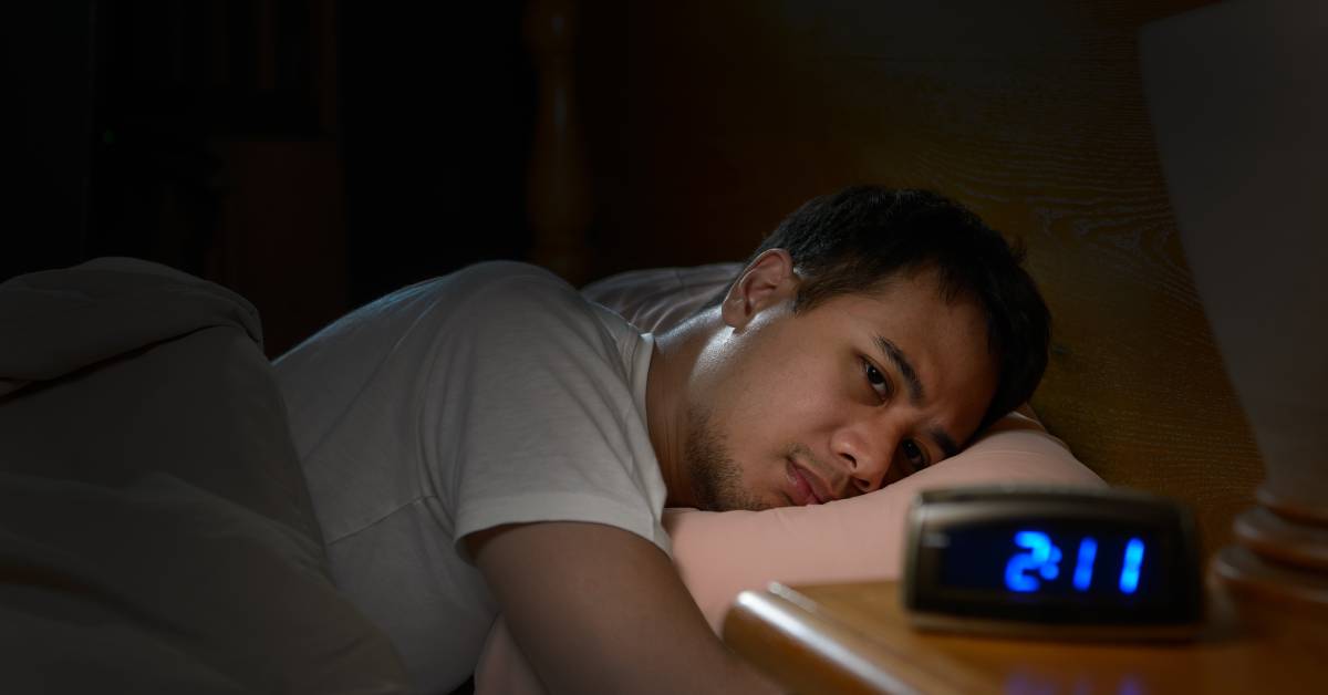 A man with insomnia lying in bed awake and looking at his alarm clock, which reads "2:11." His pillow is pink.