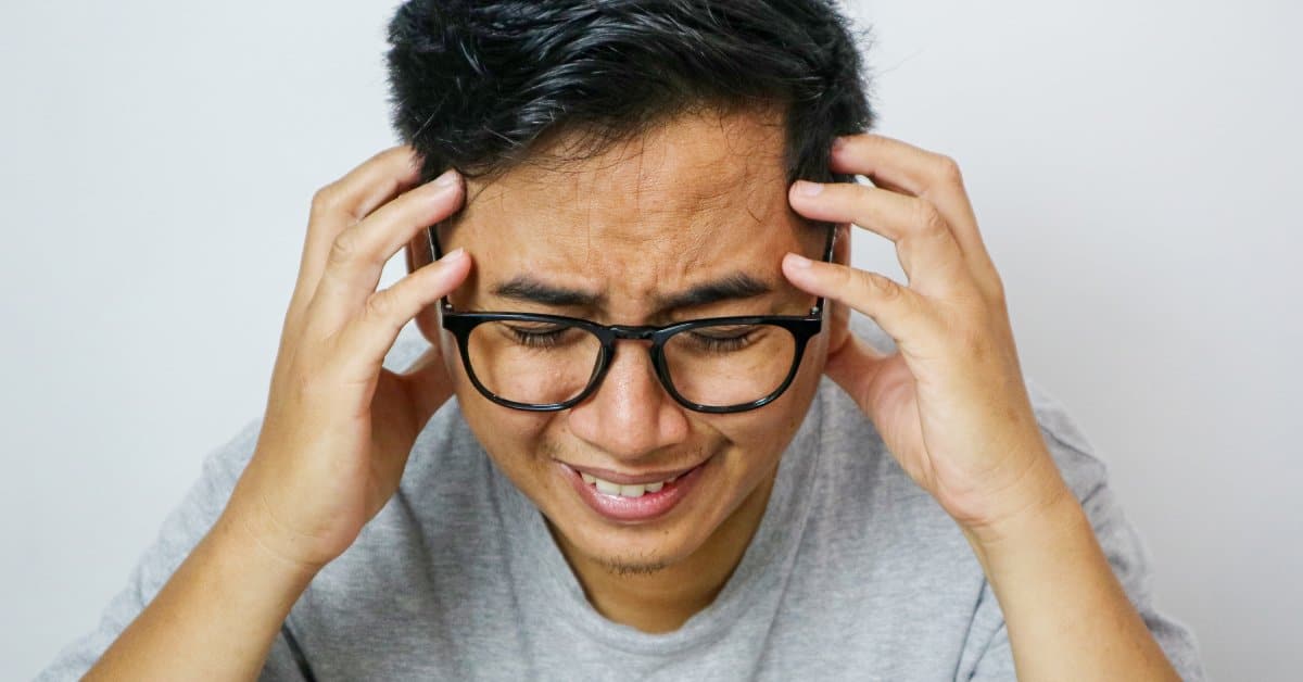 A young man looks angry and ready to cry. He wears glasses and has his fingers on each side of his forehead.