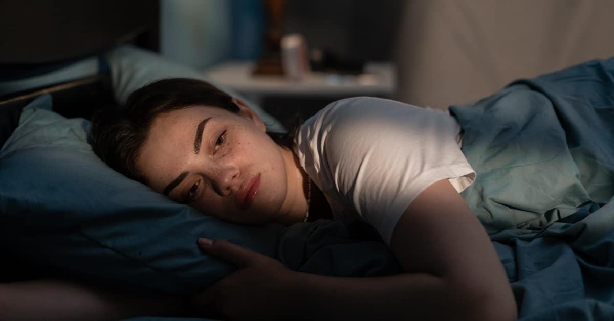 A sad-looking woman lies awake in her bed during the night. It is dark in her room except for a lamp.