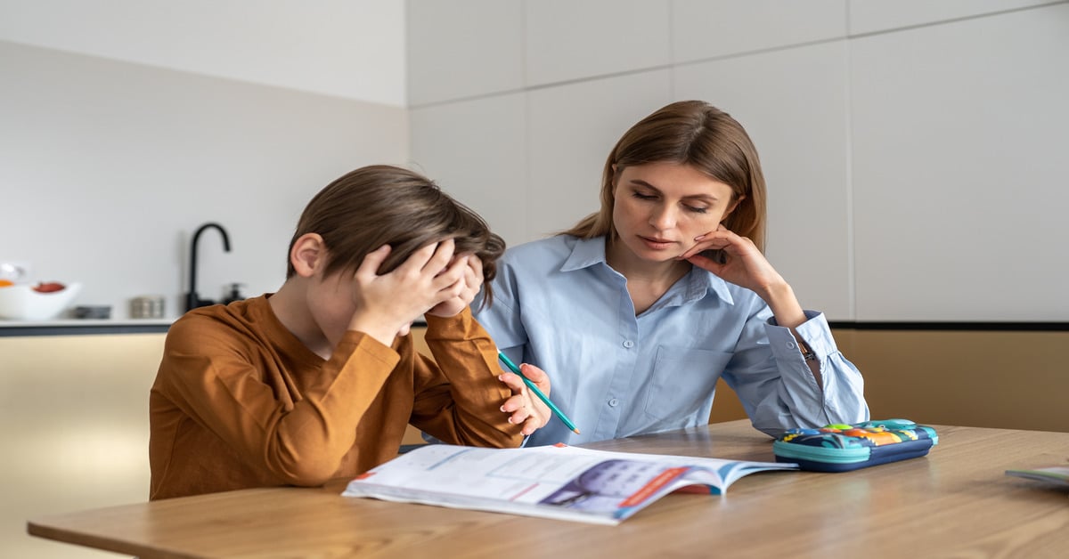 A boy puts his head in his hands, looking frustrated while trying to do homework. His mom sits next to him.