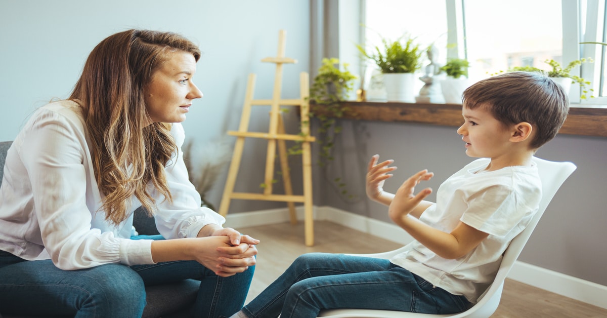 A child psychiatrist listens to a young boy as he explains something in her office. Plants sit on the windowsill behind them.