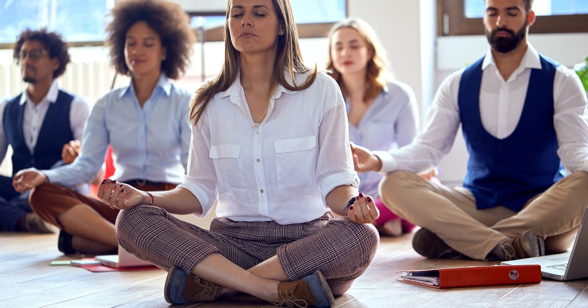A group of people practices meditation on the floor in a yoga class. Some have laptops in front of them.