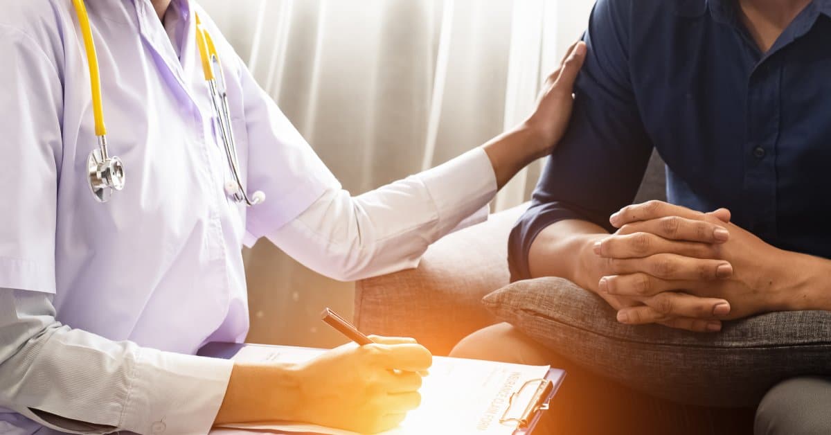 A mid-level view of a psychiatrist wearing a stethoscope and putting a supportive hand on the arm of a patient.