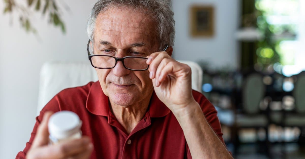 An older man sits in a bright room and looks at a prescription bottle through his glasses. He holds the pill bottle in his hand.