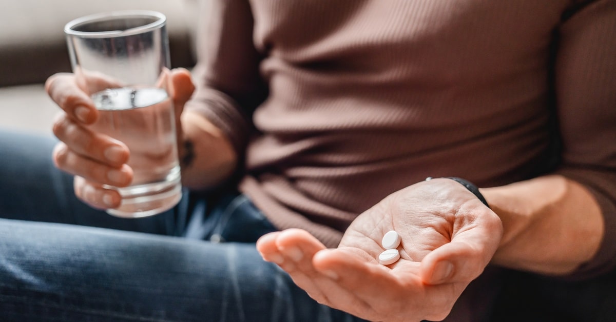 A close-up of a person's hand holding two pills. They have a glass of water in their other hand and are sitting.