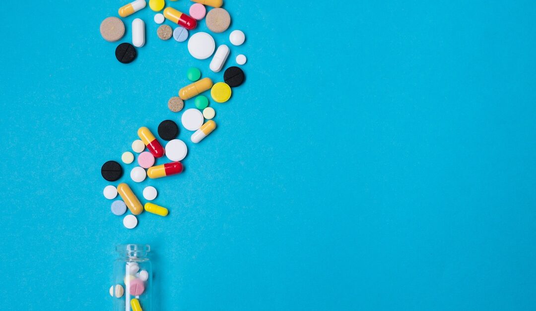 Various multicolored pills and a bottle are spilled on a blue surface, forming the shape of a question mark.