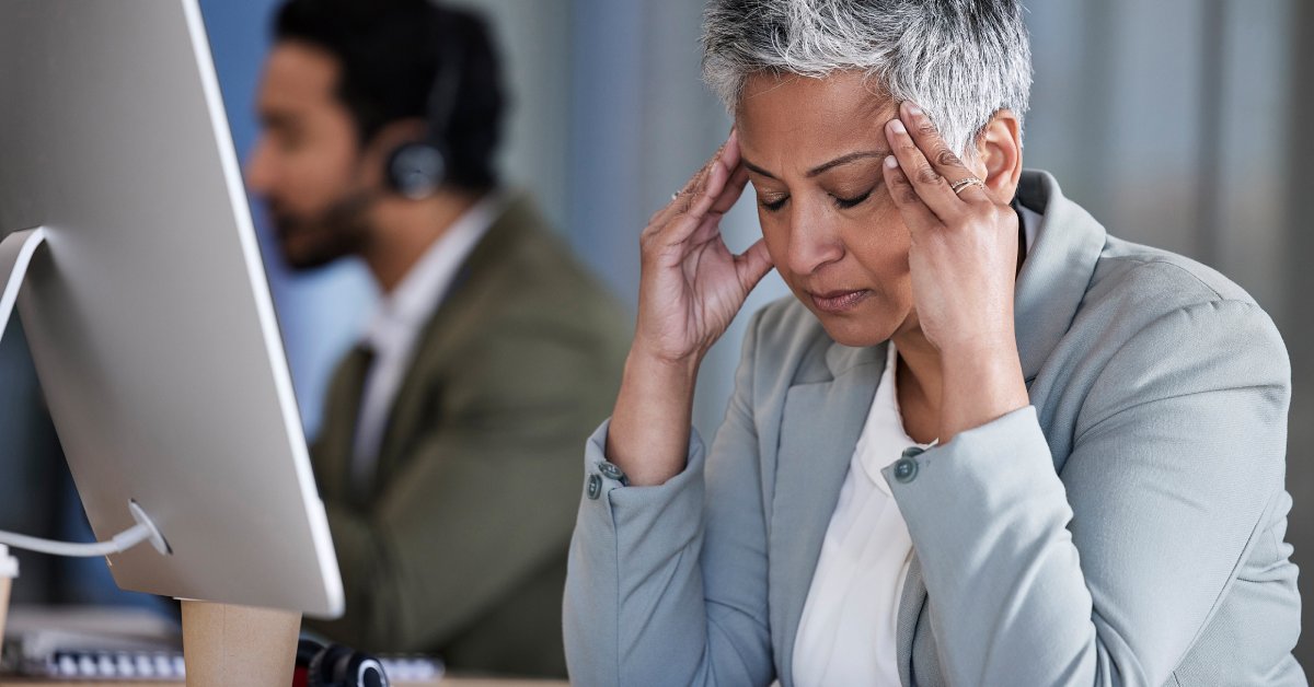 A woman rubs the sides of her forehead while looking stressed. She sits at a desk with a computer in an office.