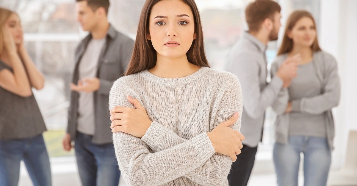 A woman stands in a room, looking nervous, while other people stand around her, talking. She wears a gray sweater.