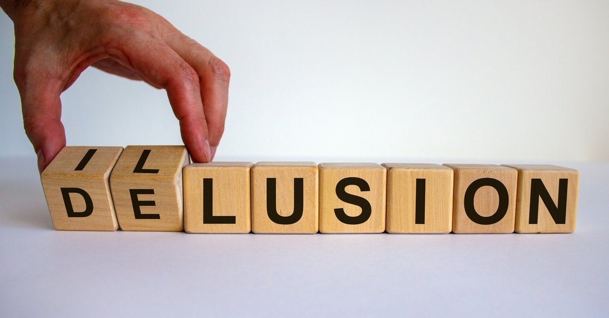 A hand turns letter blocks so that they spell out the word "Delusion." They sit on a white background.