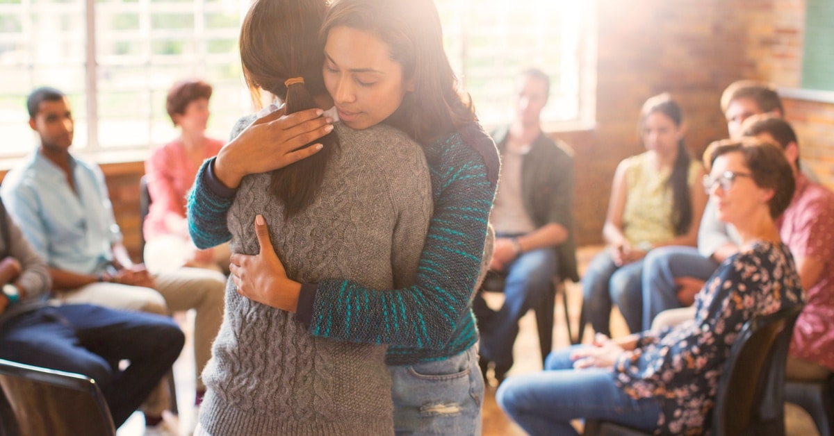 Two women hug while surrounded by people at a group therapy session. The session room has brick walls.