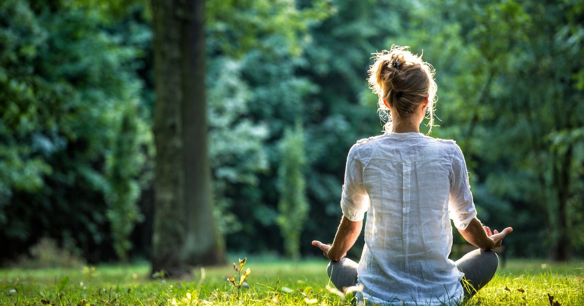 A woman meditates while sitting in a field. The sun shines on the grass, and several trees are in the background.