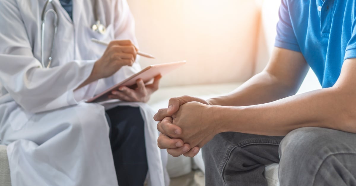 A person wearing a white lab coat is sitting across from another person. The one in the white coat is holding a clipboard.