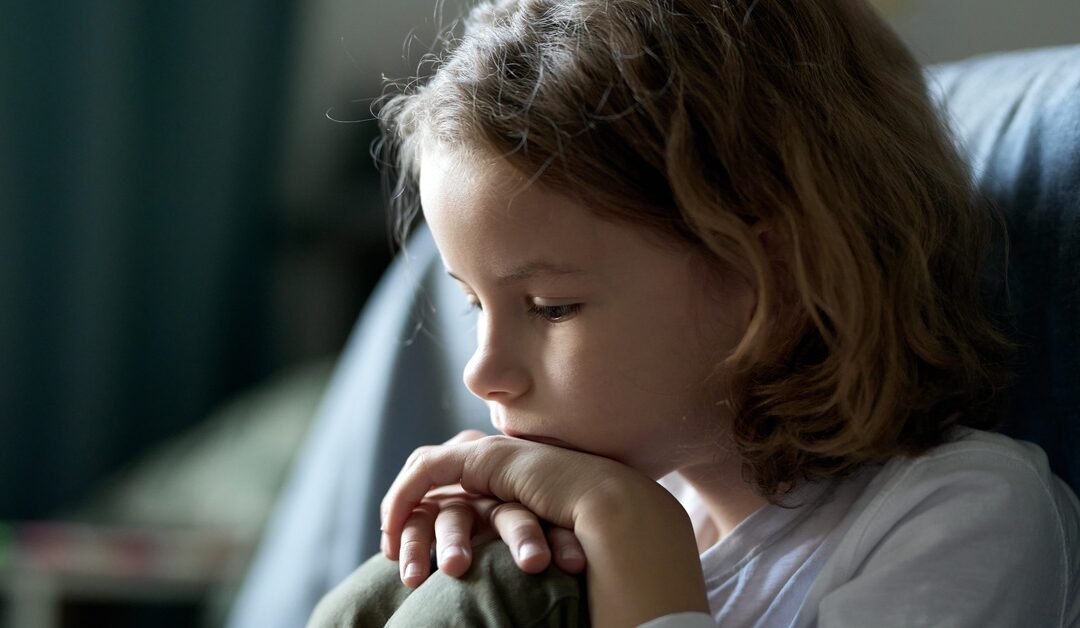 A young child sits in his room, looking sad. He has his chin in his hands, which are on top of his knees.