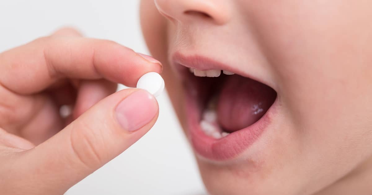 A close-up of an adult's hand placing a white pill into a child's open mouth. The background behind them is white.