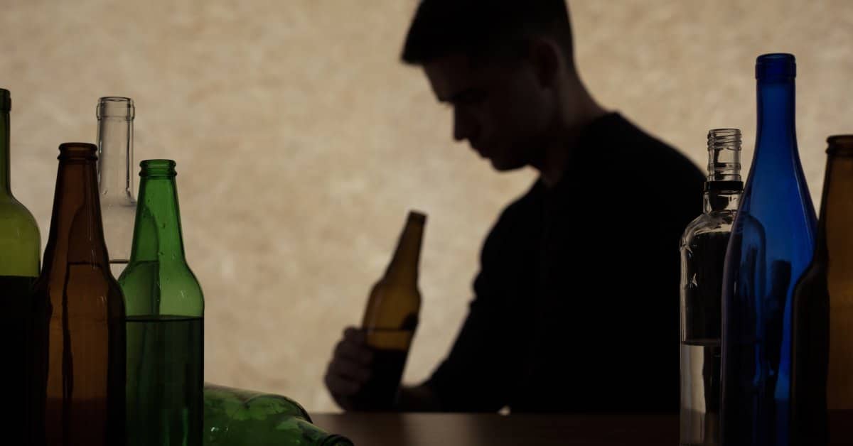 An adolescent boy looking at a half-empty bottle of beer. Several bottles of alcohol sit in the foreground on a table.