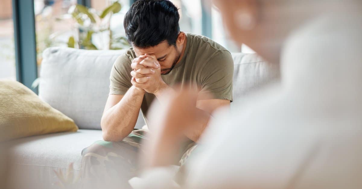 A man dressed in a green shirt and camouflage pants sitting on a couch with his head in his hands, while a person listens.