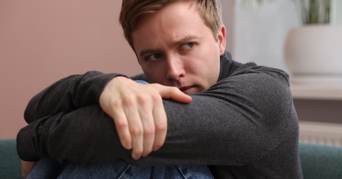A man sits with his knees to his chest and his arms on his legs. He has a frightened look on his face.