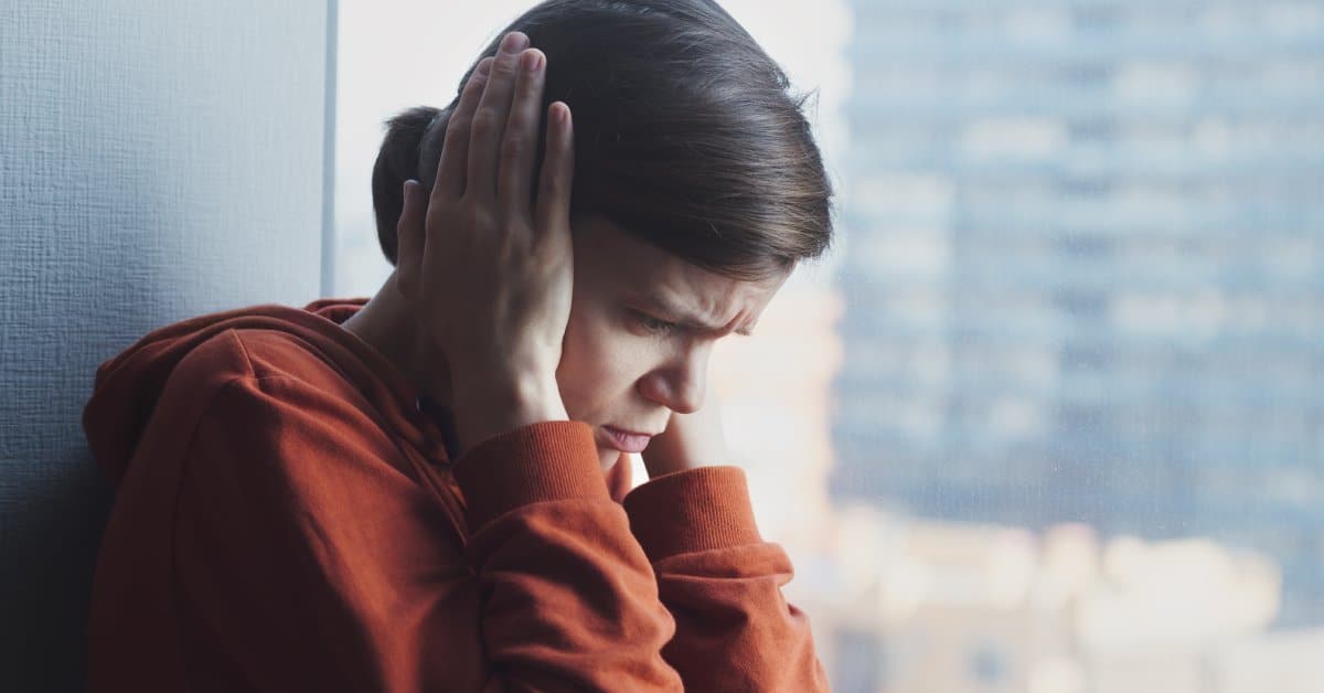 A woman with a sad look on her face stands in a corner, covering her ears. The window behind her shows a building.
