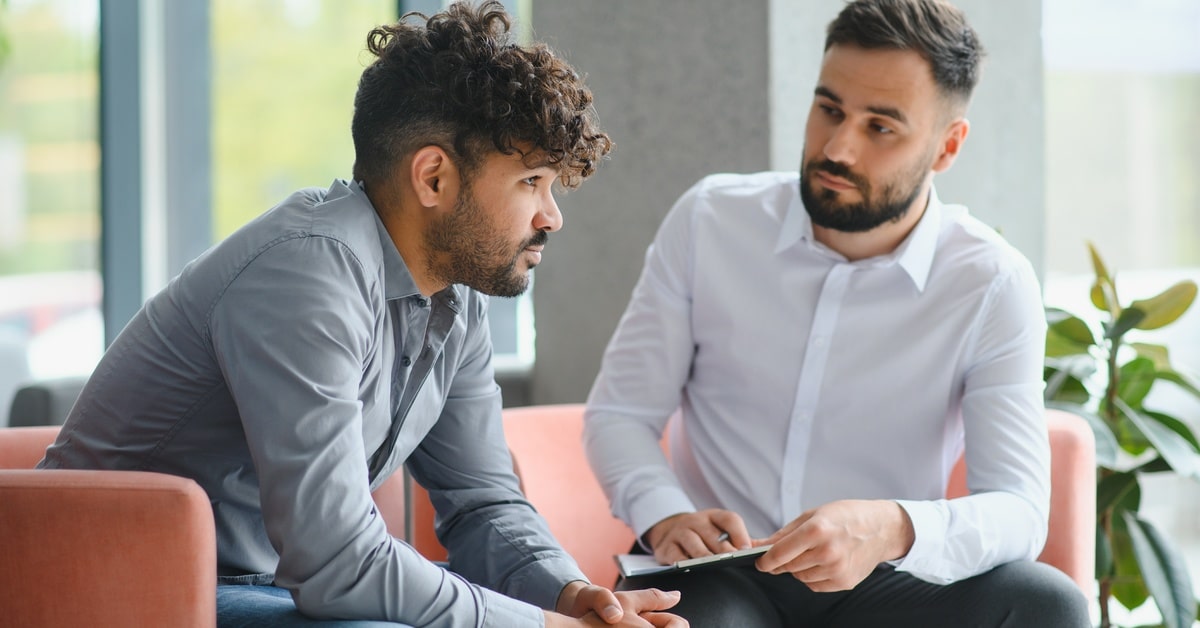 A sad-looking man sits on a couch next to a psychiatrist in his office. The latter person listens and makes notes.