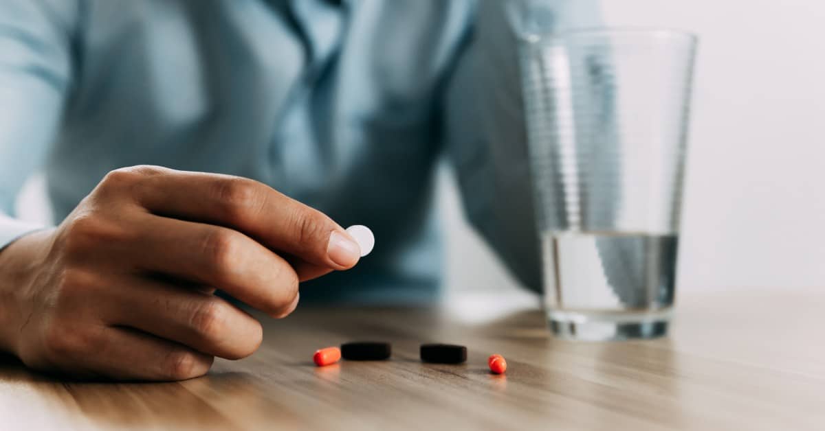 Close-up of a man's hand picking up a white pill off a table. Next to his hand is a full glass of water.