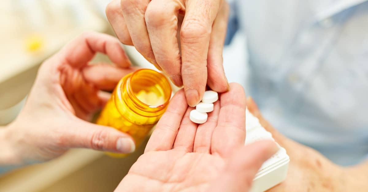 Close-up of a person's hand holding pills in their palm. Another person reaches over with their hand to pick one up.